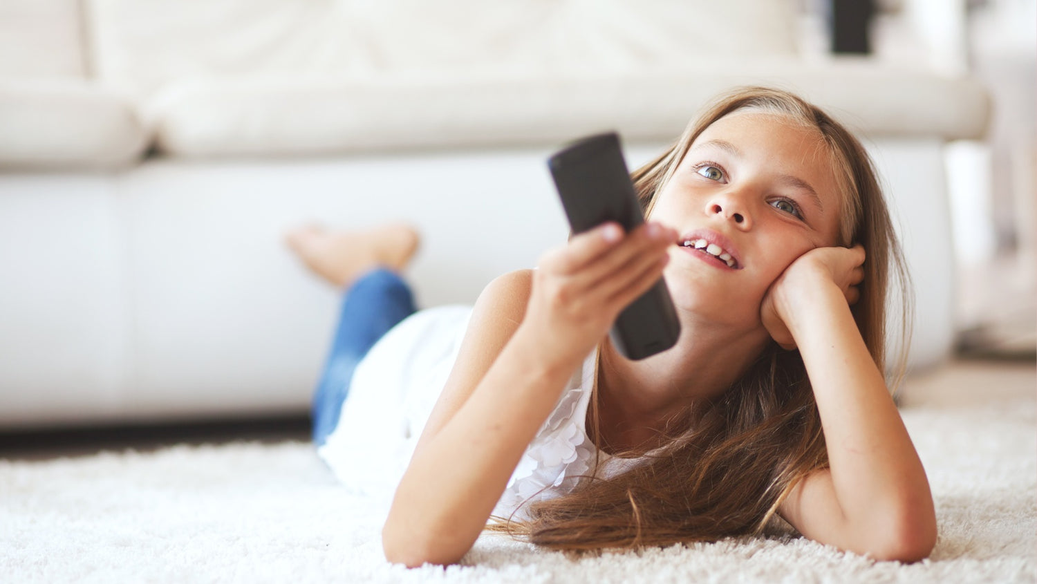 Child laying on her chest on top of a rug. She is resting her head in one palm, with a TV remote in her other hand. She is presumably facing a TV.