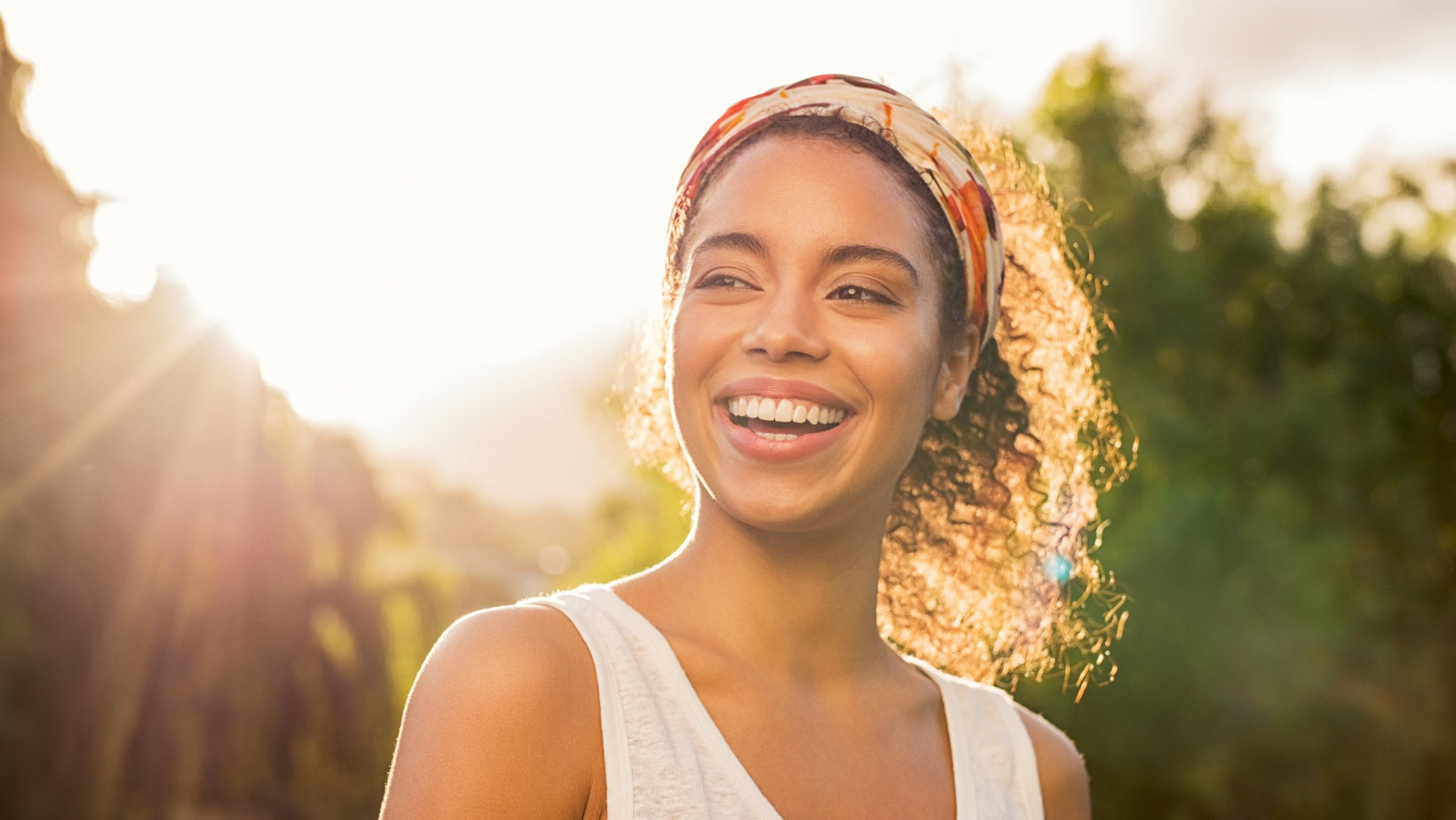 Woman standing outside, close up of face and upper body. She is smiling while looking off to her side. The sun is shining in the background, creating a lens flare.