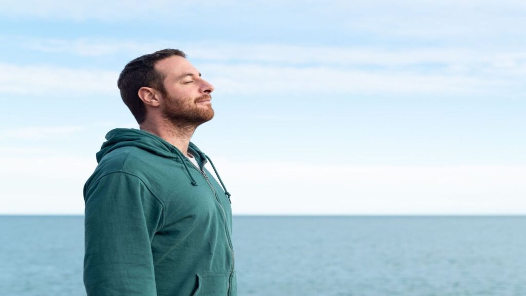 Nose breathing vs mouth breathing featured image - man standing breathing through his nose, expressing comfort, ocean backdrop.